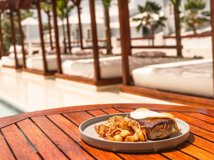 Una mesa de madera con un plato de comida, una piscina y un pabellón al fondo.