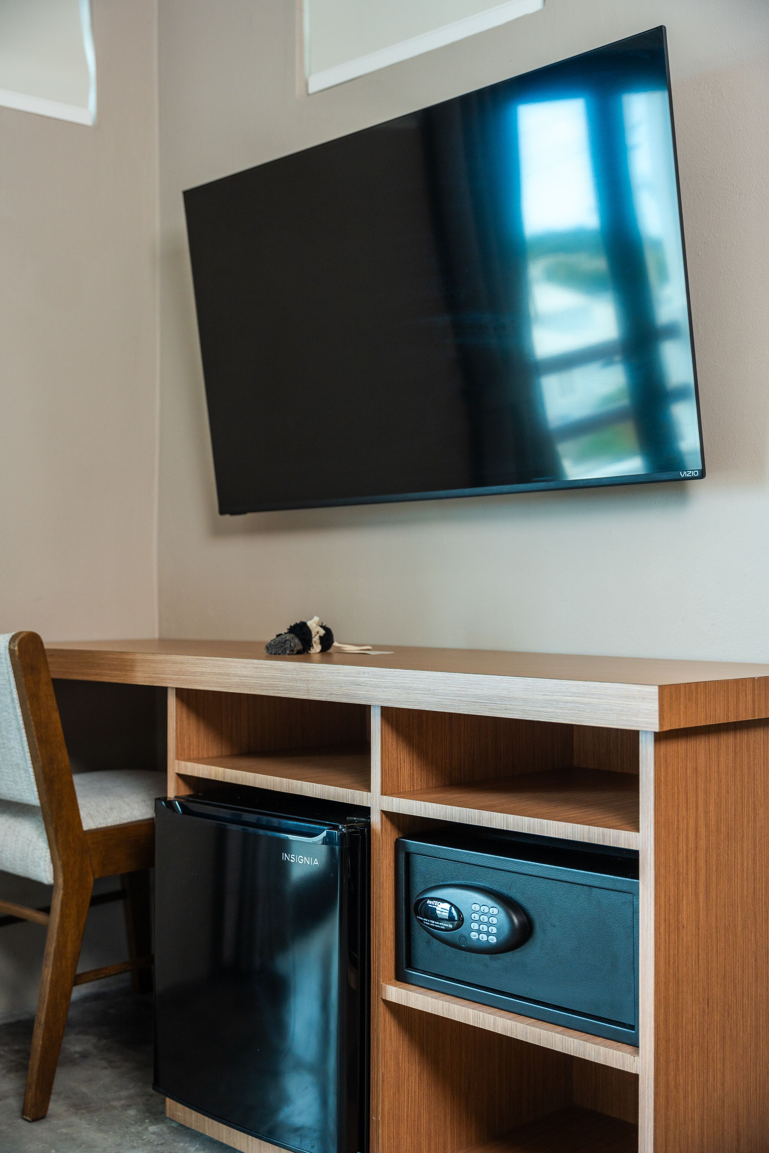 Habitación compacta con un escritorio de madera, un refrigerador negro, una caja fuerte y un televisor de pantalla plana montado en la pared.