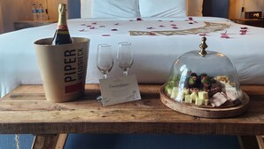 A hotel room with a bed, a bedside table with a bottle of champagne, two glasses, a card, and a cheese platter under a glass dome. - Parguera Plaza Hotel - Adults Only (Lajas)