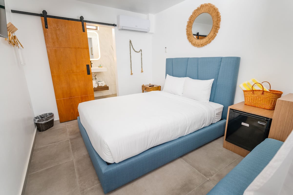Ein Schlafzimmer mit einem Bett, einem Nachttisch, einem kleinen Holfnachtschränkchen, einem Korbwägelchen und einem Spiegel an der Wand.