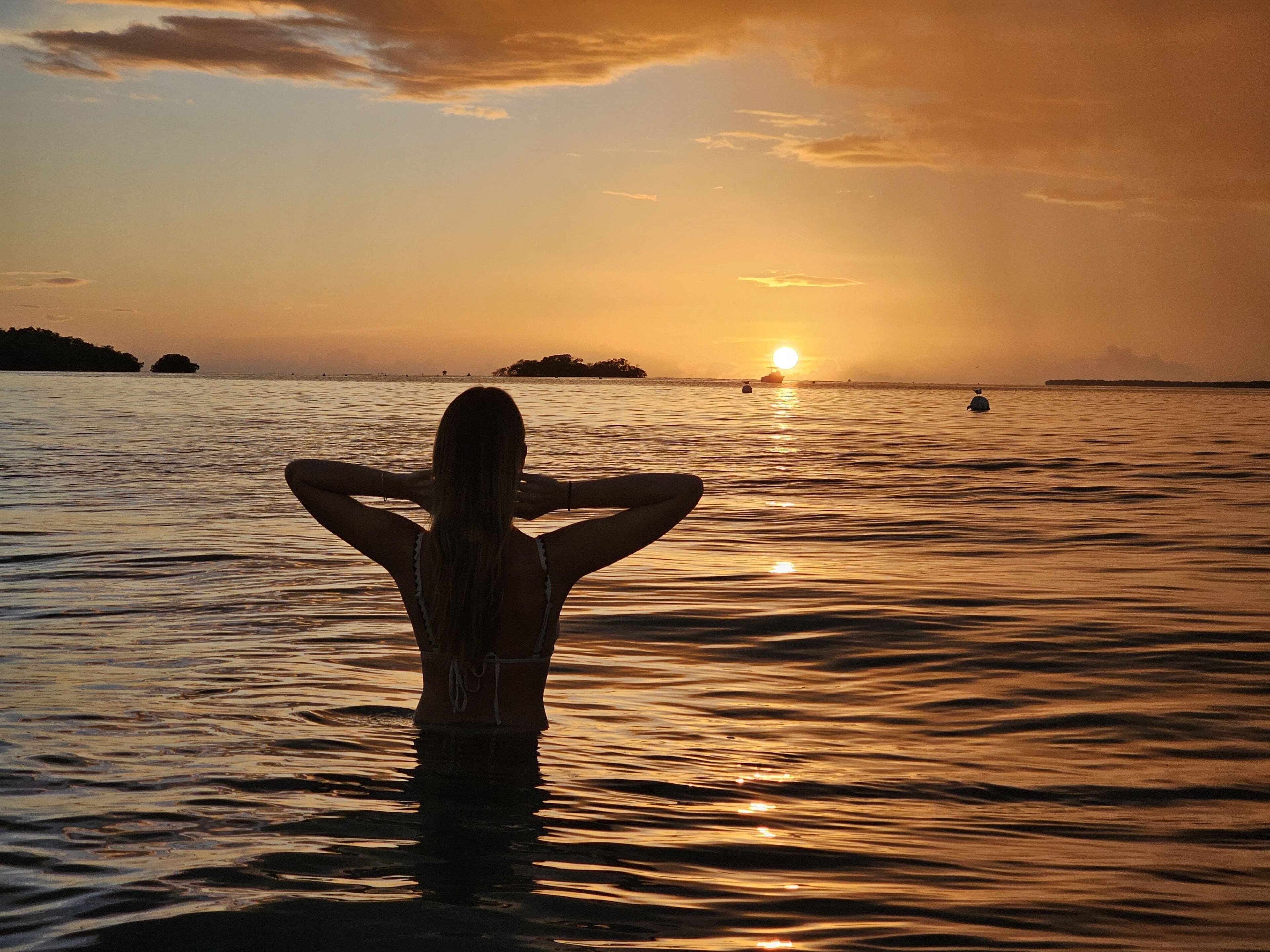 A person standing in water facing a sunset with a distant island and a boat.