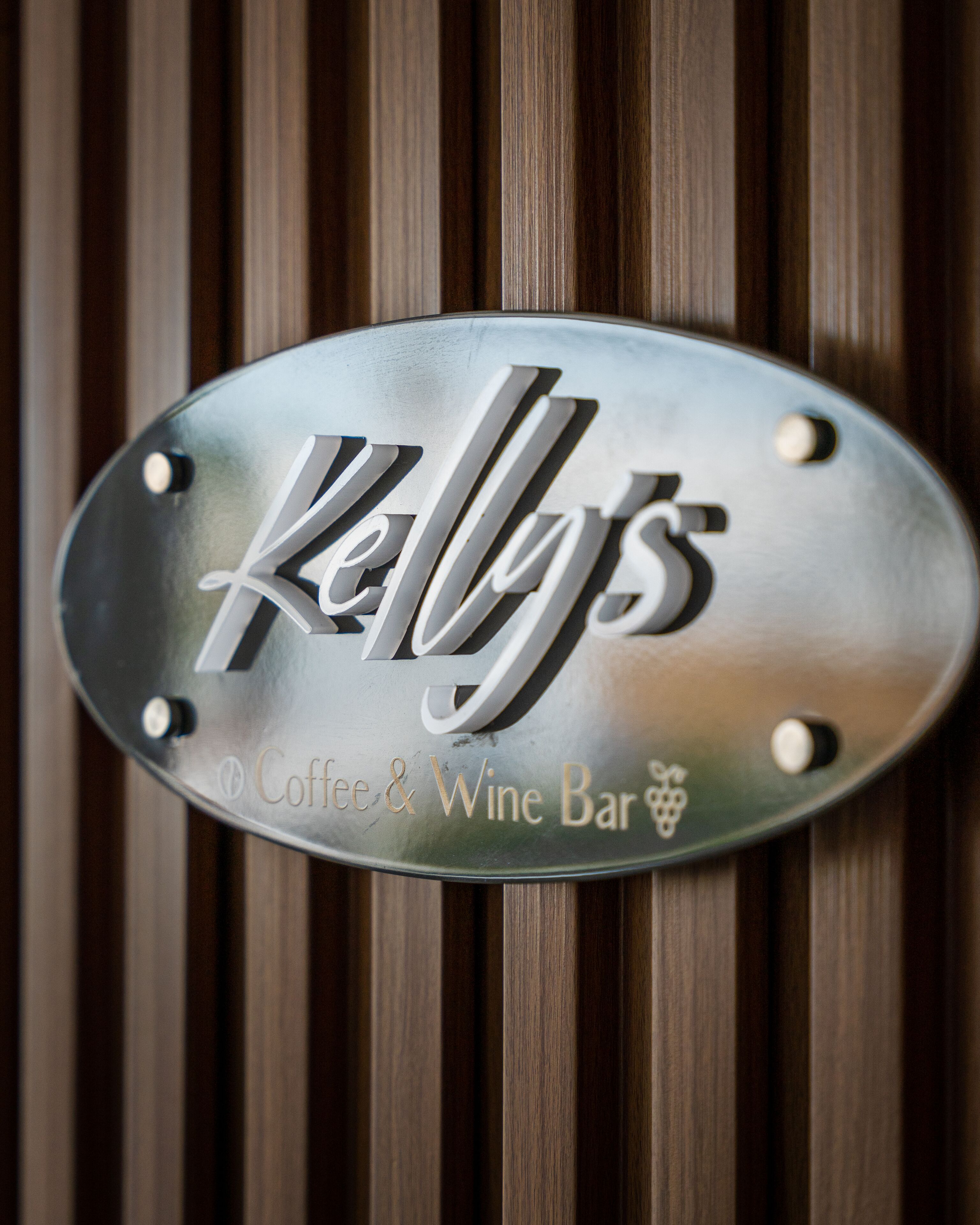 Une enseigne métallique portant l’inscription « Kelly’s Coffee & Wine Bar » est fixée sur une surface en bois.