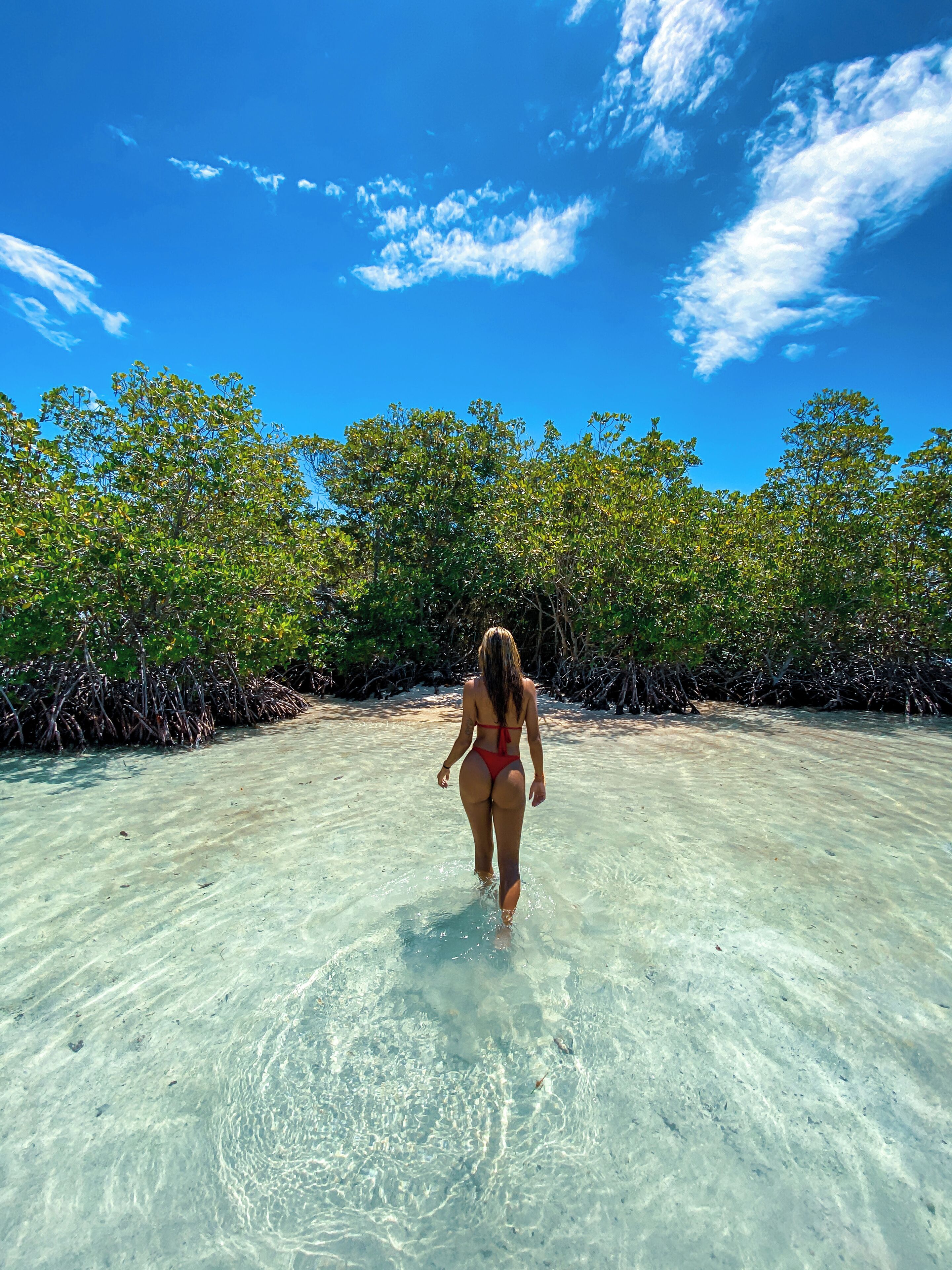 A woman walking on a beach with clear water and mangrove trees in the background.