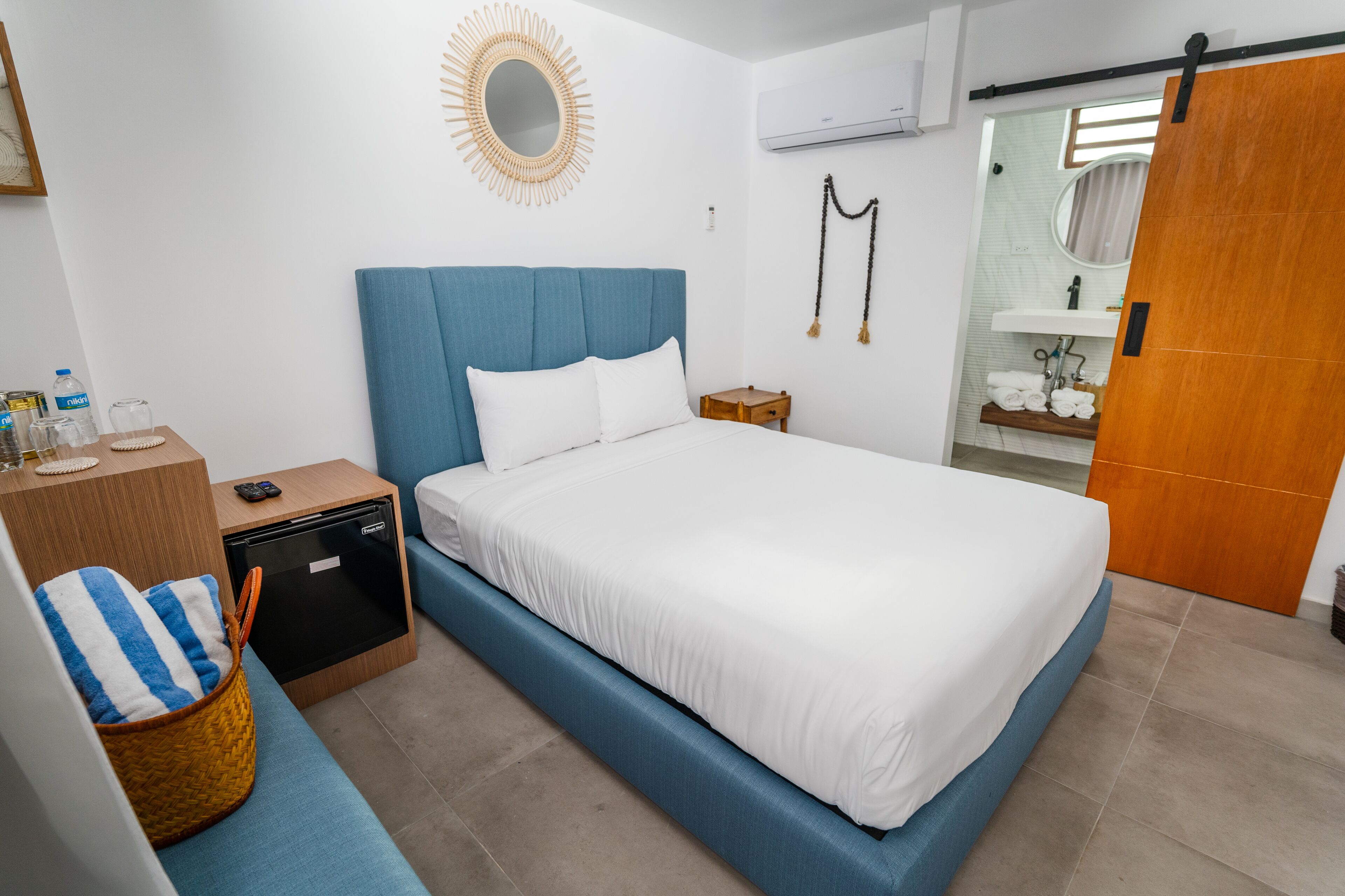 Une chambre d’hôtel moderne avec un grand lit, une petite table de chevet, une commode, un panier en osier, un miroir et une salle de bain équipée d’un lavabo et d’un porte-serviettes.