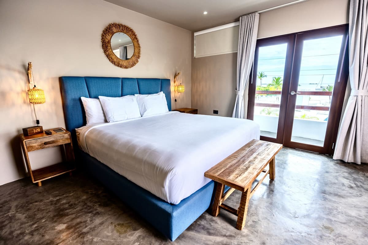 Ein Hotelzimmer mit einem großen Bett, einem Nachttisch aus Holz, einer Nachttischlampe, einem Spiegel und Blick ins Freie durch eine Glastür.