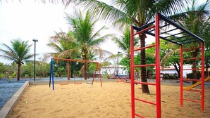 Ein Spielplatz mit roter und blauer Spielausstattung, umgeben von Sand und Palmen.