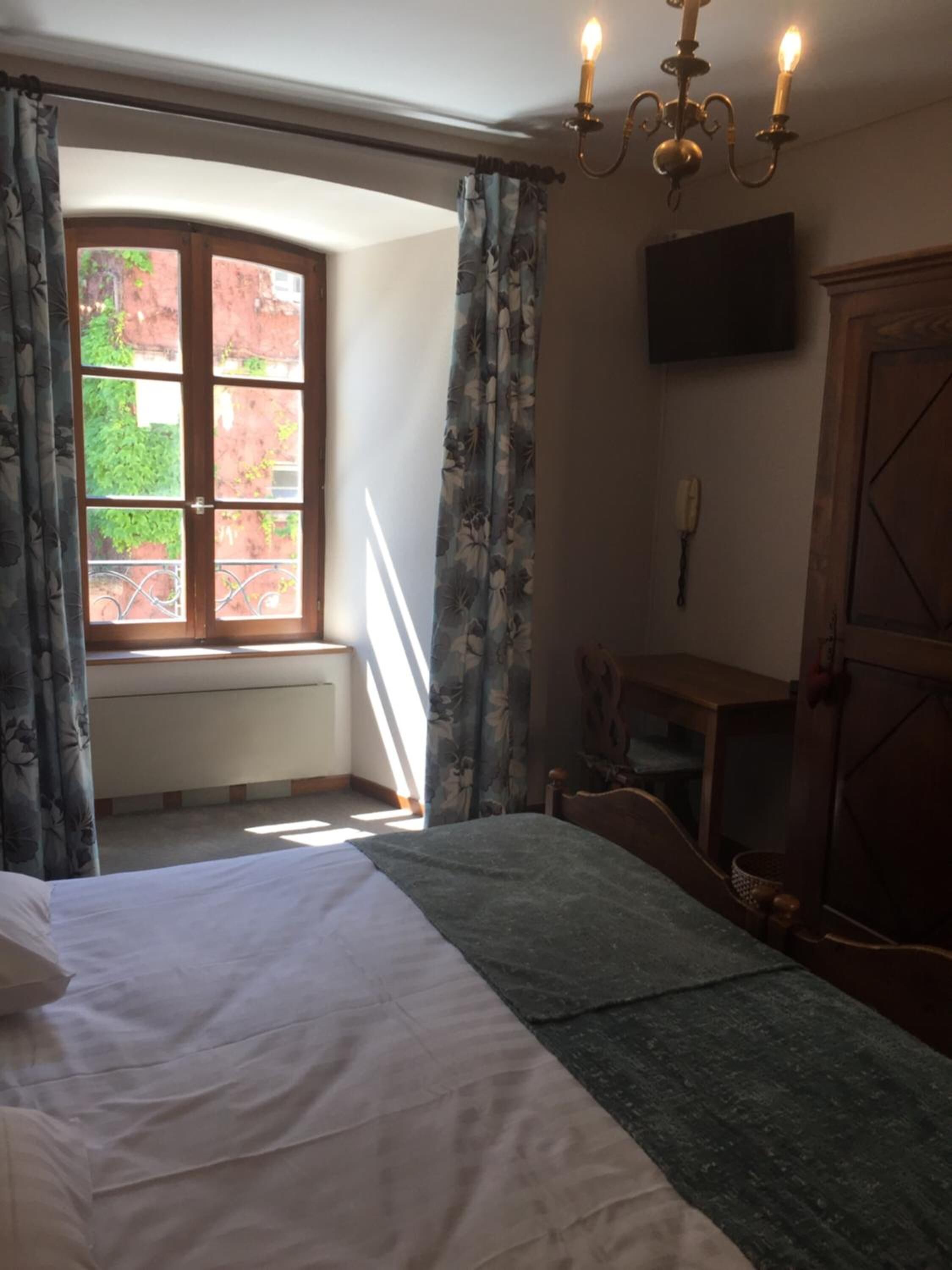 A bedroom with a bed, a window with curtains, a chandelier, a television, and a wooden cabinet.