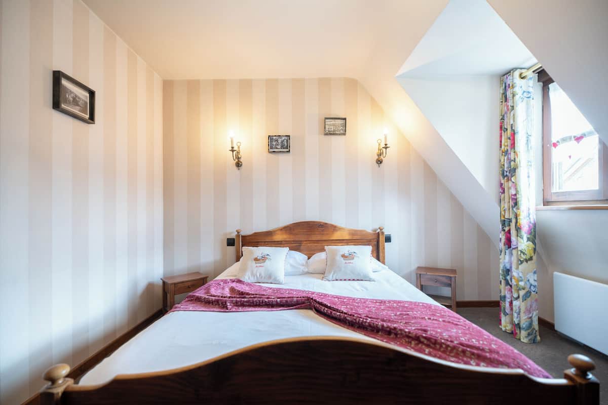 A bedroom with a sloped ceiling, a wooden bed, two bedside tables, and a window with a curtain.