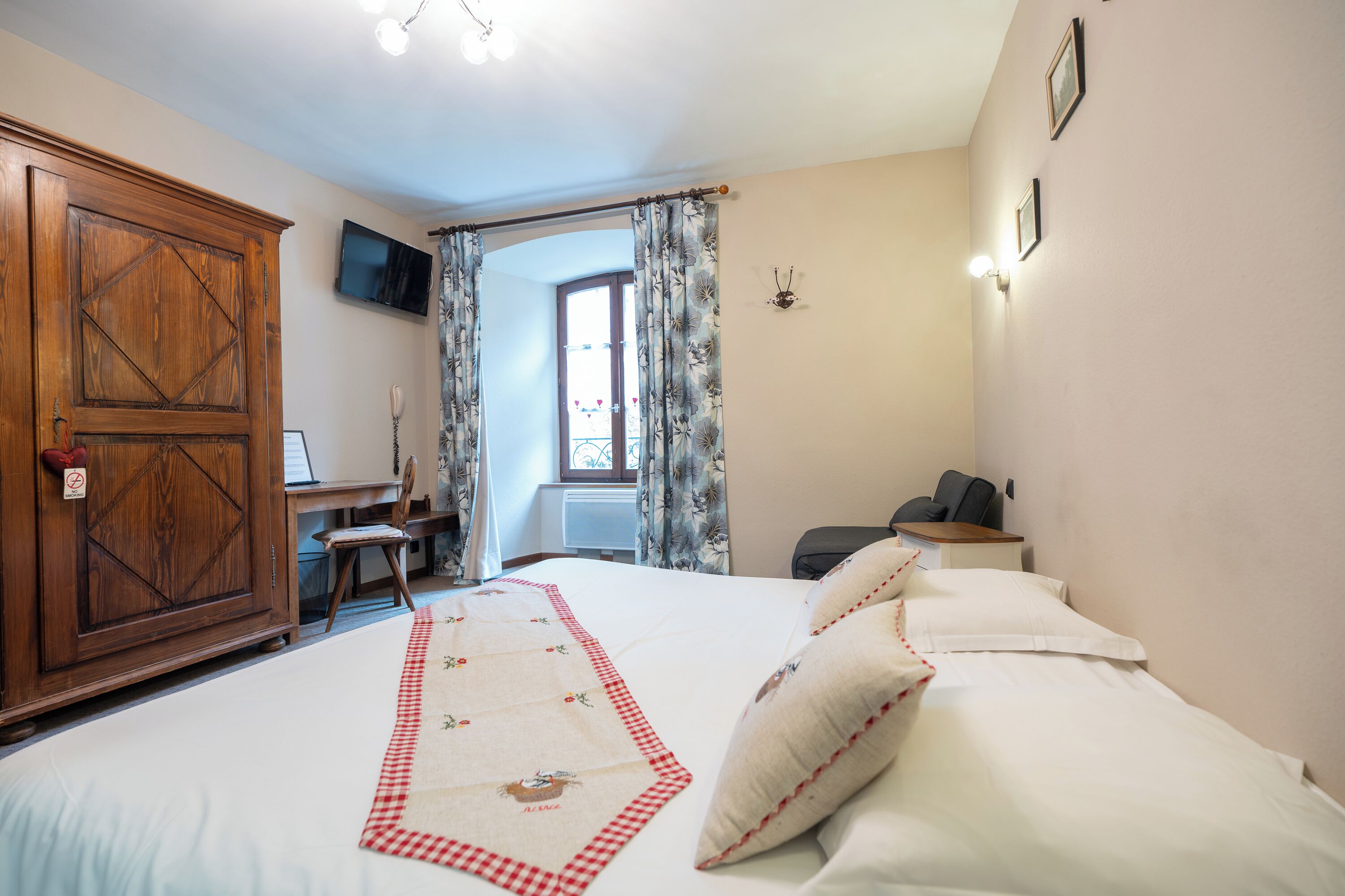 Una camera da letto con un letto grande, un armadio in legno, una scrivania con una sedia, una televisione a parete e una finestra con tende.