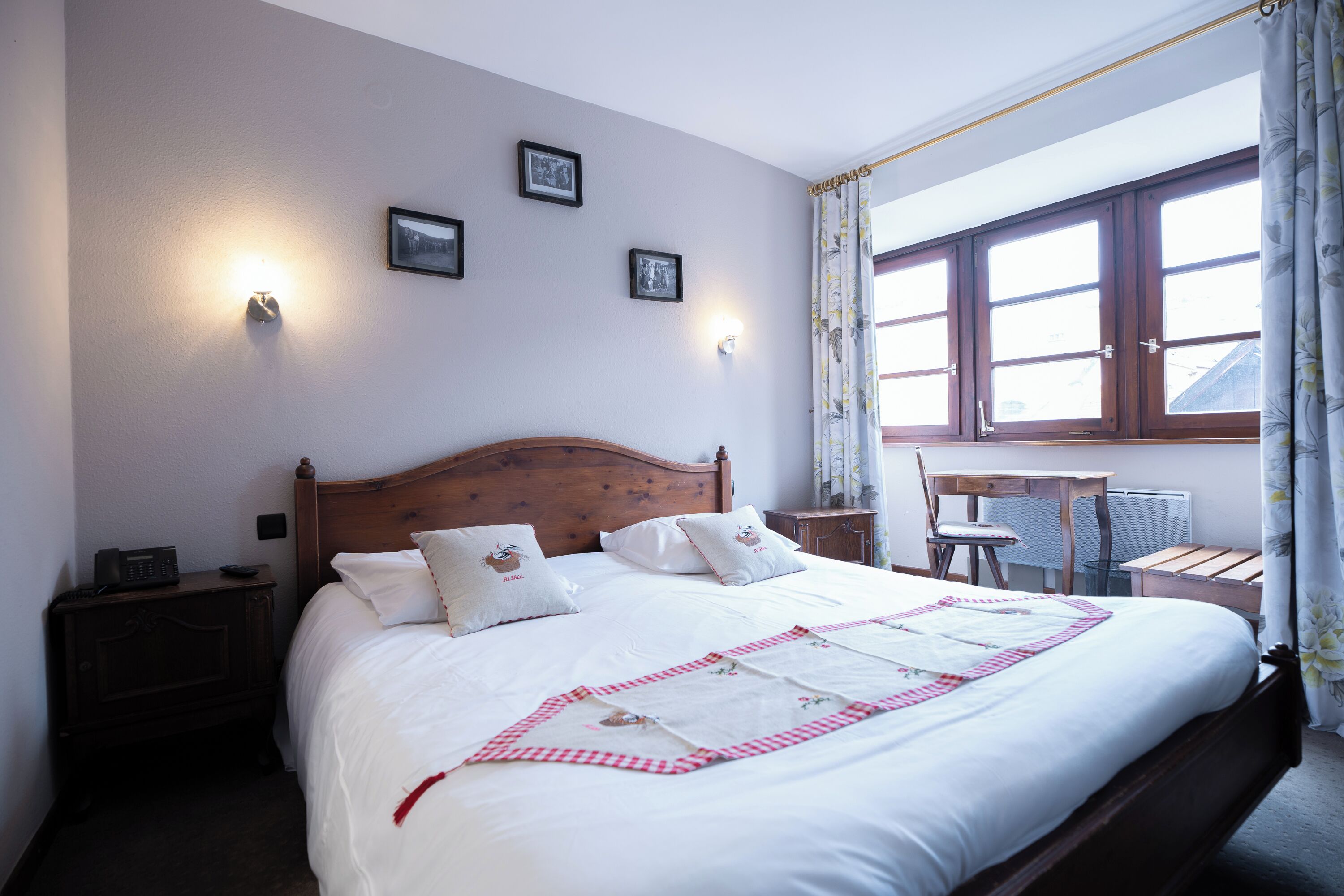 A neatly made bed with white linens and a red and white checkered blanket, a wooden headboard, bedside tables with lamps, and a window with curtains.