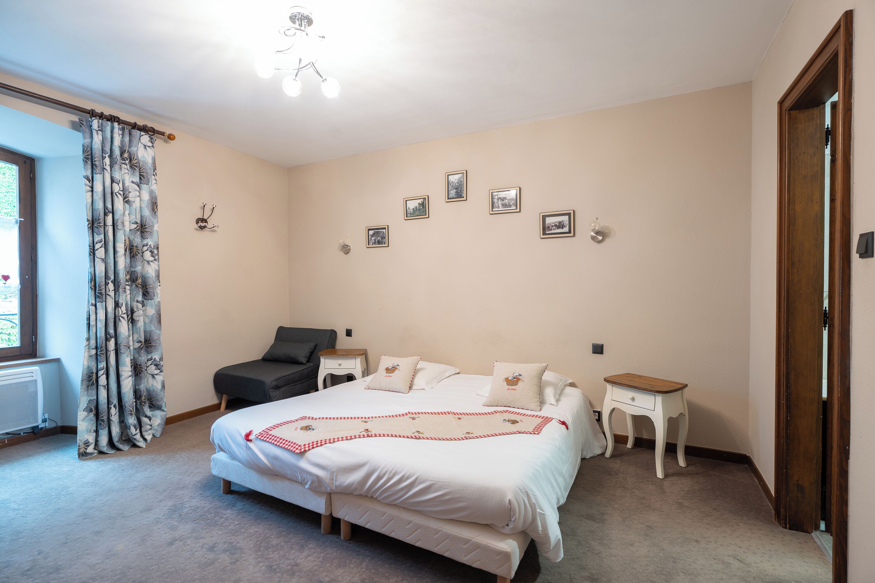 A bedroom with a bed, bedside tables, a chair, a window with curtains, and wall-mounted pictures.