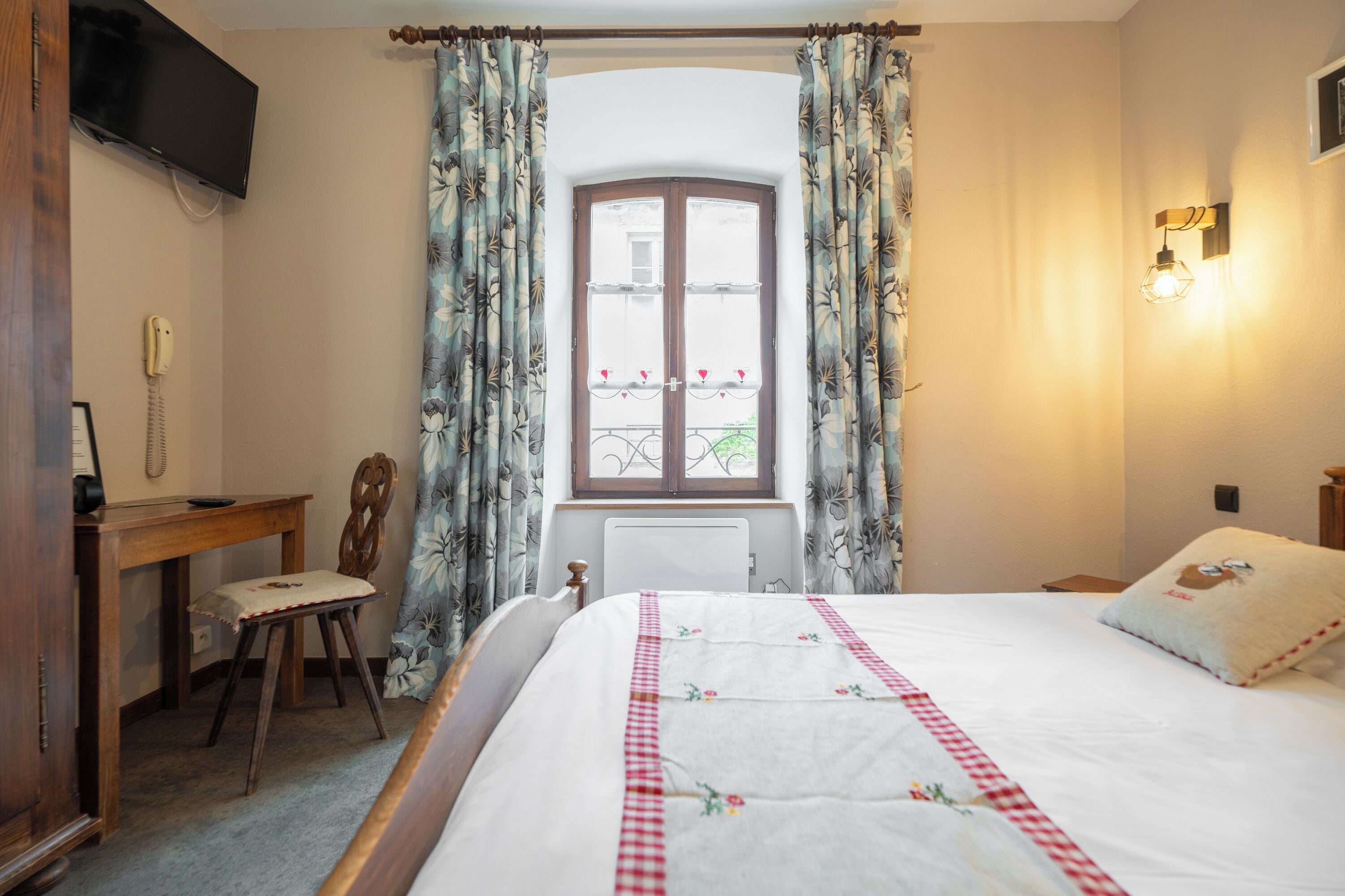 Een slaapkamer met een bed, een bureau, een stoel, een televisie en een raam met gordijnen.
