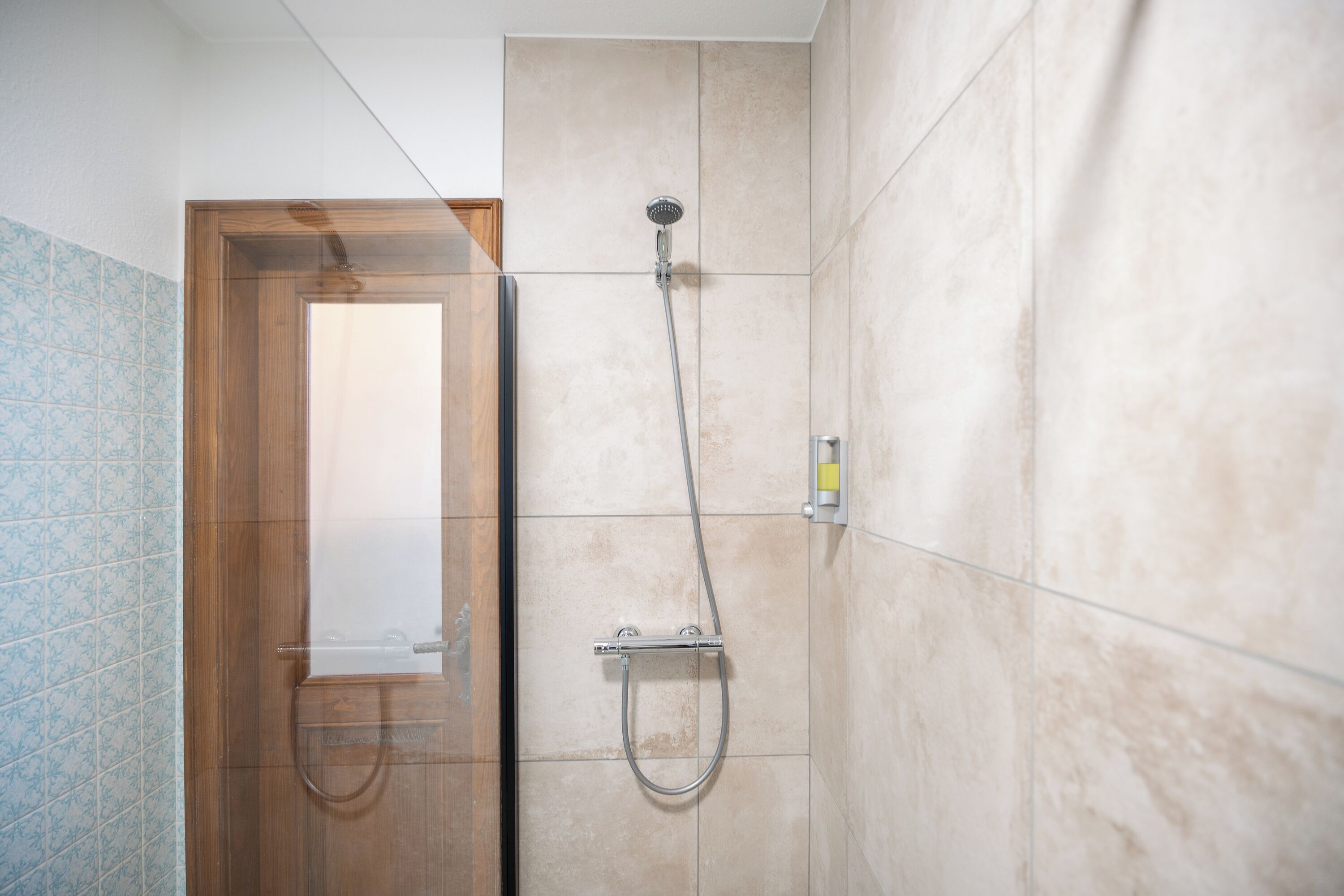 A shower area with a handheld showerhead and a wall-mounted faucet, a wooden door, and a tiled wall with a blue pattern on one side.