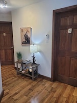 Un pasillo con piso de madera, una consola de madera con una lámpara, un cuadro enmarcado y dos puertas con números.