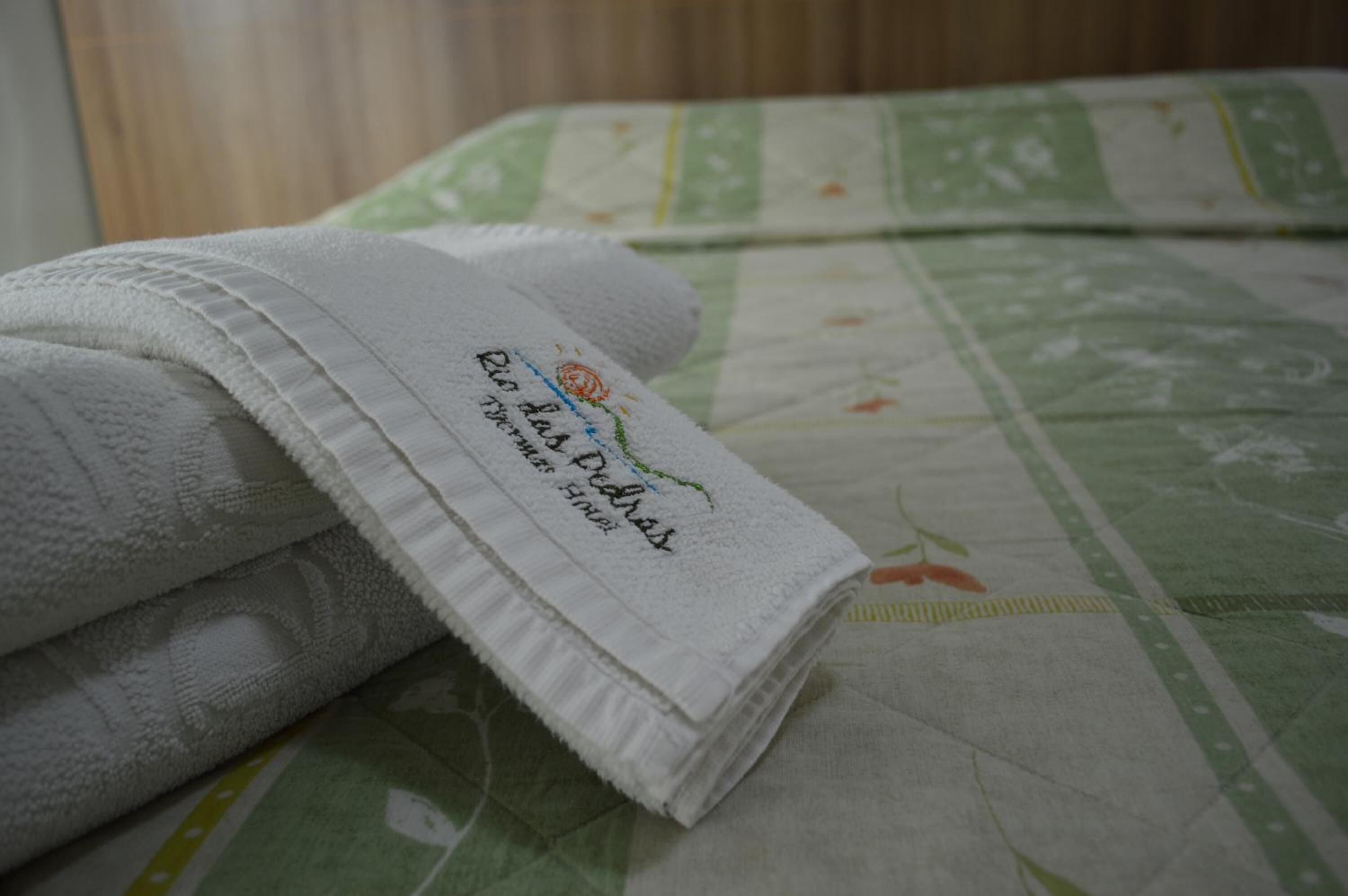 Una toalla blanca doblada con cuidado y texto bordado sobre una cama con colcha de flores verde y blanca.