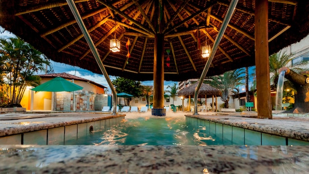 A pool area with a thatched roof structure, lounge chairs, and a stone wall. - Rio das Pedras Thermas Hotel (Caldas Novas)