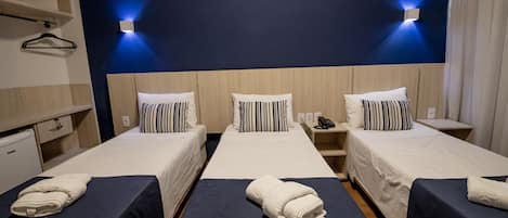 Habitación de hotel con tres camas individuales, cada una con una colcha azul y almohadas blancas. Hay dos lámparas de pared en la pared azul oscuro y un cabecero de madera.