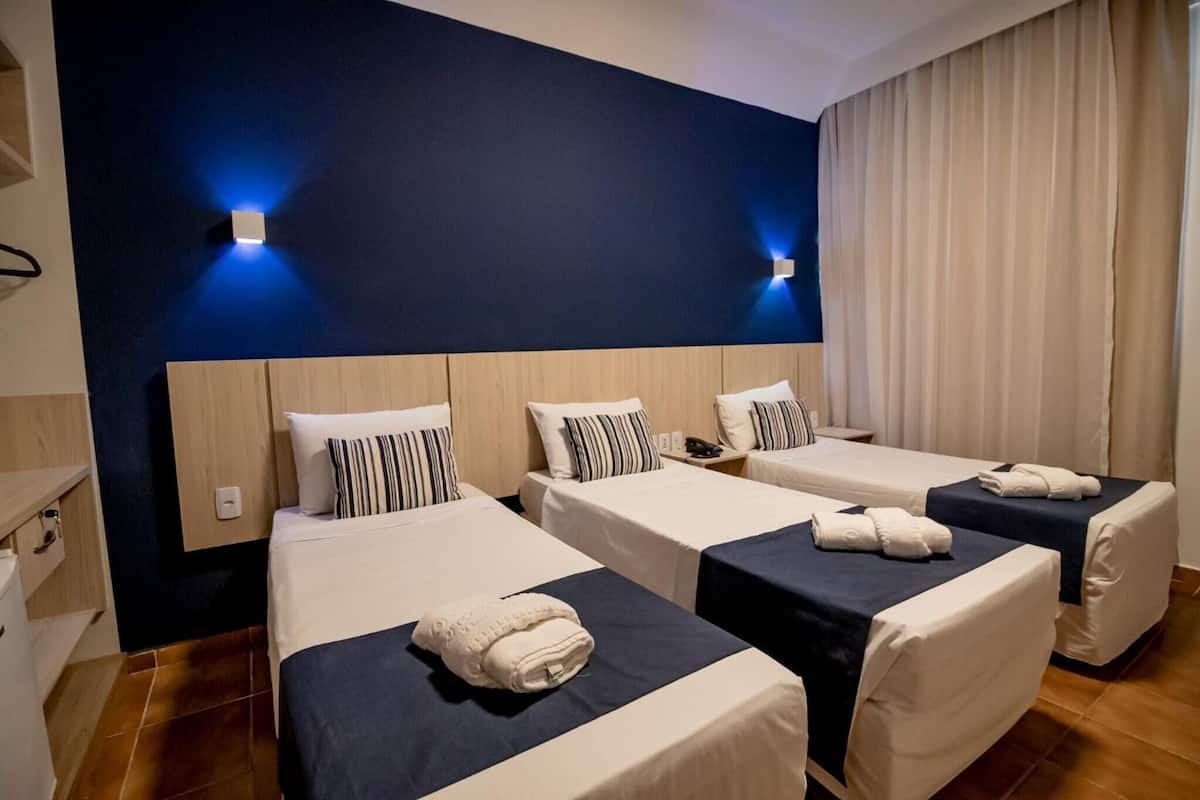 A hotel room with three single beds, each with a blue bedspread and white pillows. There are two wall-mounted light fixtures on the dark blue wall.