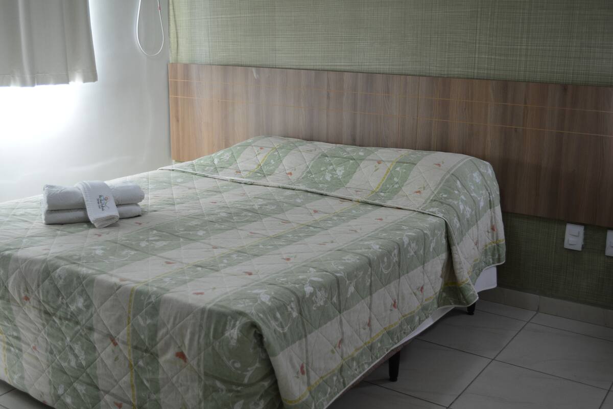 A neatly made bed with a quilted green and white pattern, a folded towel on the left side, and a wooden headboard.