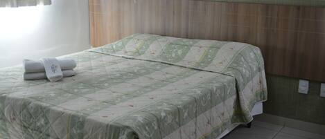 Una cama bien tendida con un estampado verde y blanco acolchado, una toalla doblada a la izquierda y un cabecero de madera.