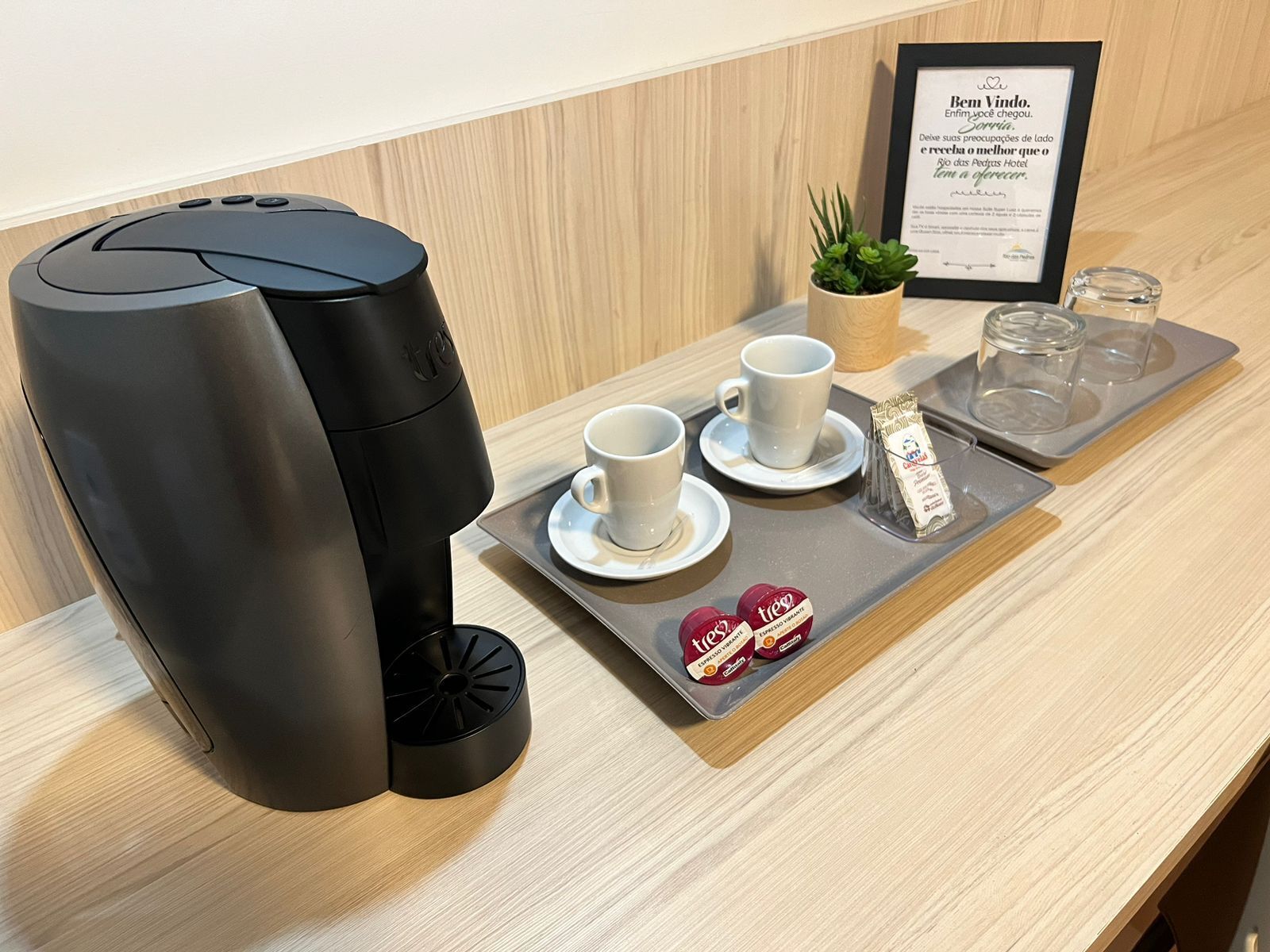 A coffee machine, two white cups, a small plant, a framed sign, and a tray with a coffee pod and a glass.