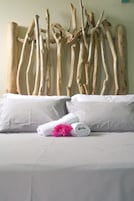 Una cama con sábana blanca, dos almohadas y una toalla enrollada con una flor rosa.