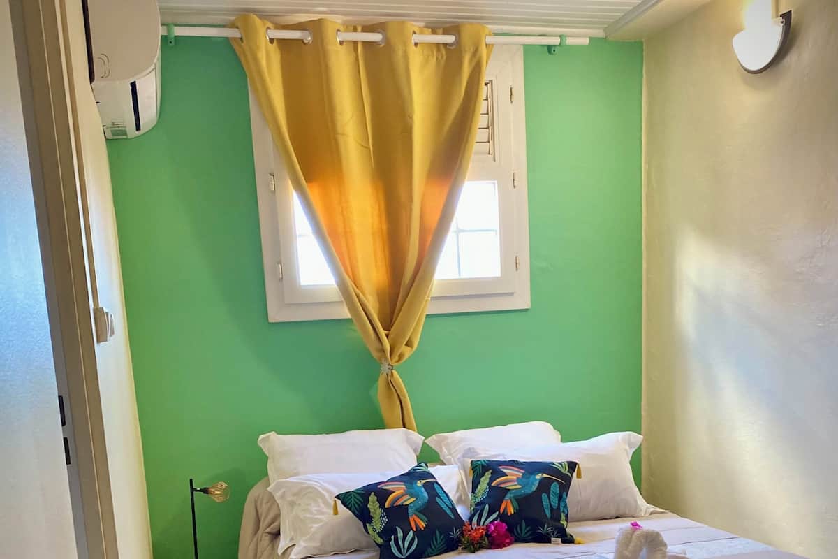 Une chambre à coucher avec un lit, des oreillers, un petit tabouret et une fenêtre avec des rideaux.
