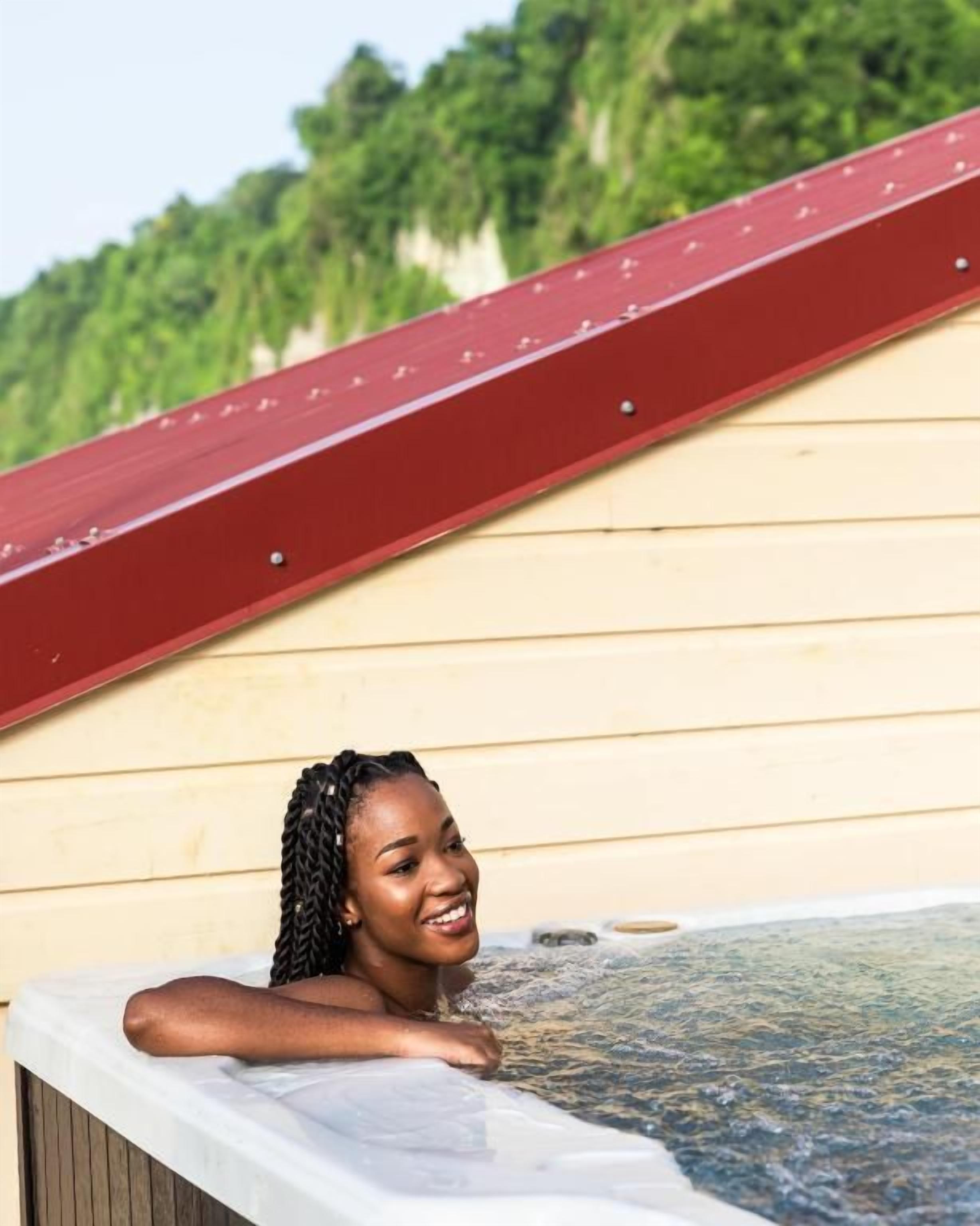 A person relaxing in a hot tub with a wooden wall and a red roof in the background.