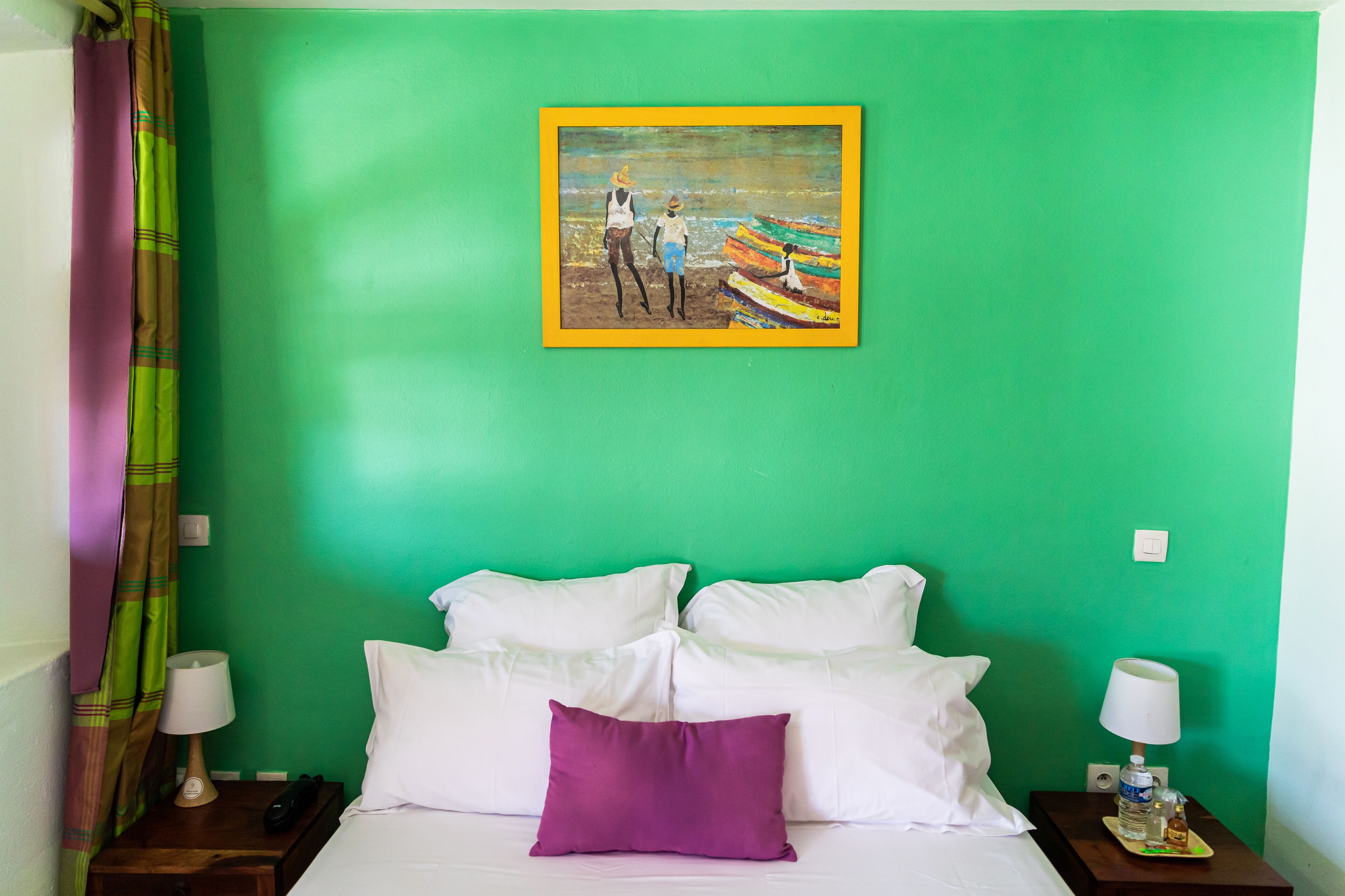 Habitación de hotel con una pared verde, una cama con almohadas blancas y púrpuras, un cuadro con gente en una playa, y una mesita de noche con una lámpara y una botella.