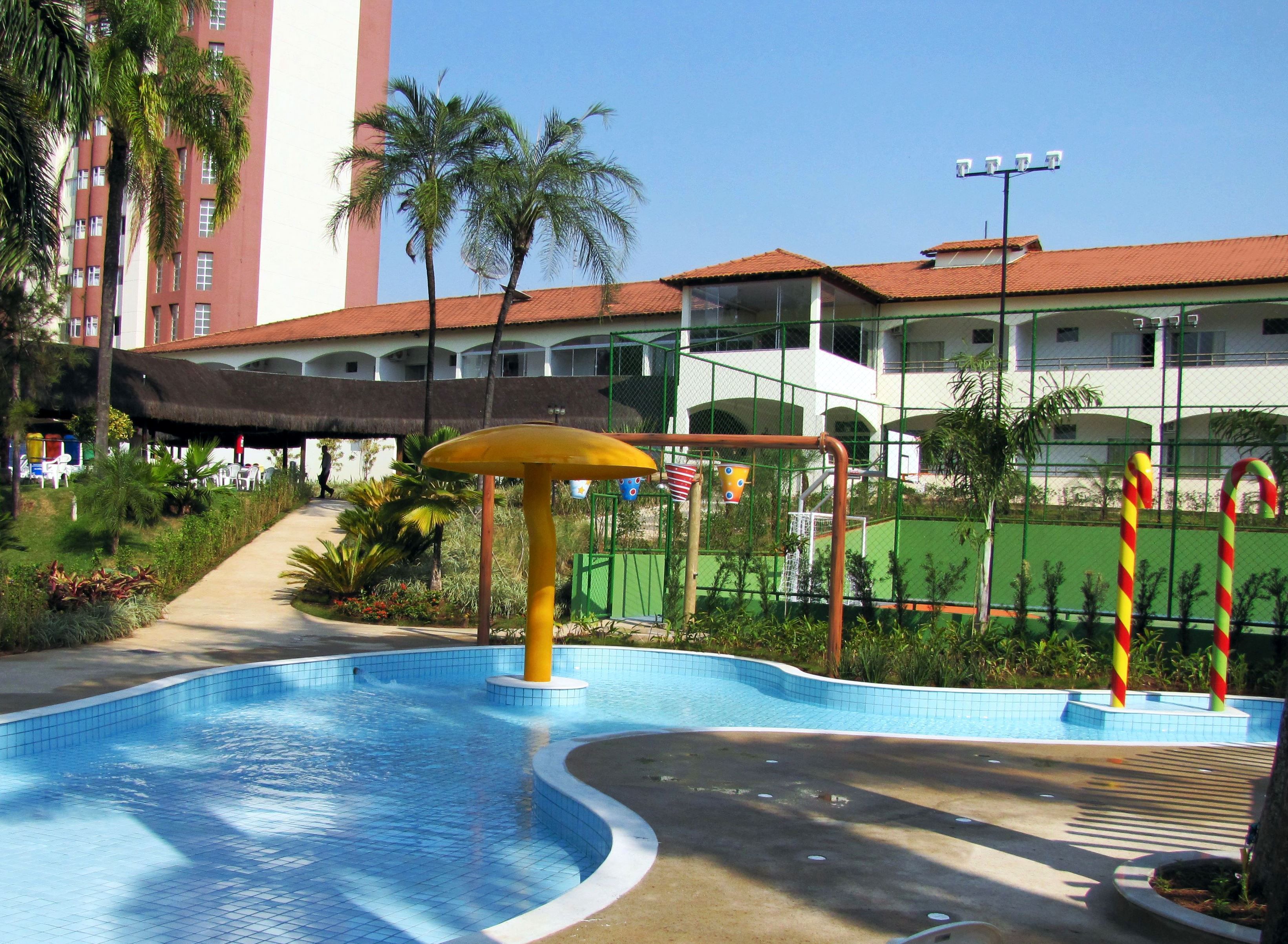 A swimming pool with a curved edge, surrounded by a paved walkway. There are palm trees and a building with a red roof in the background.