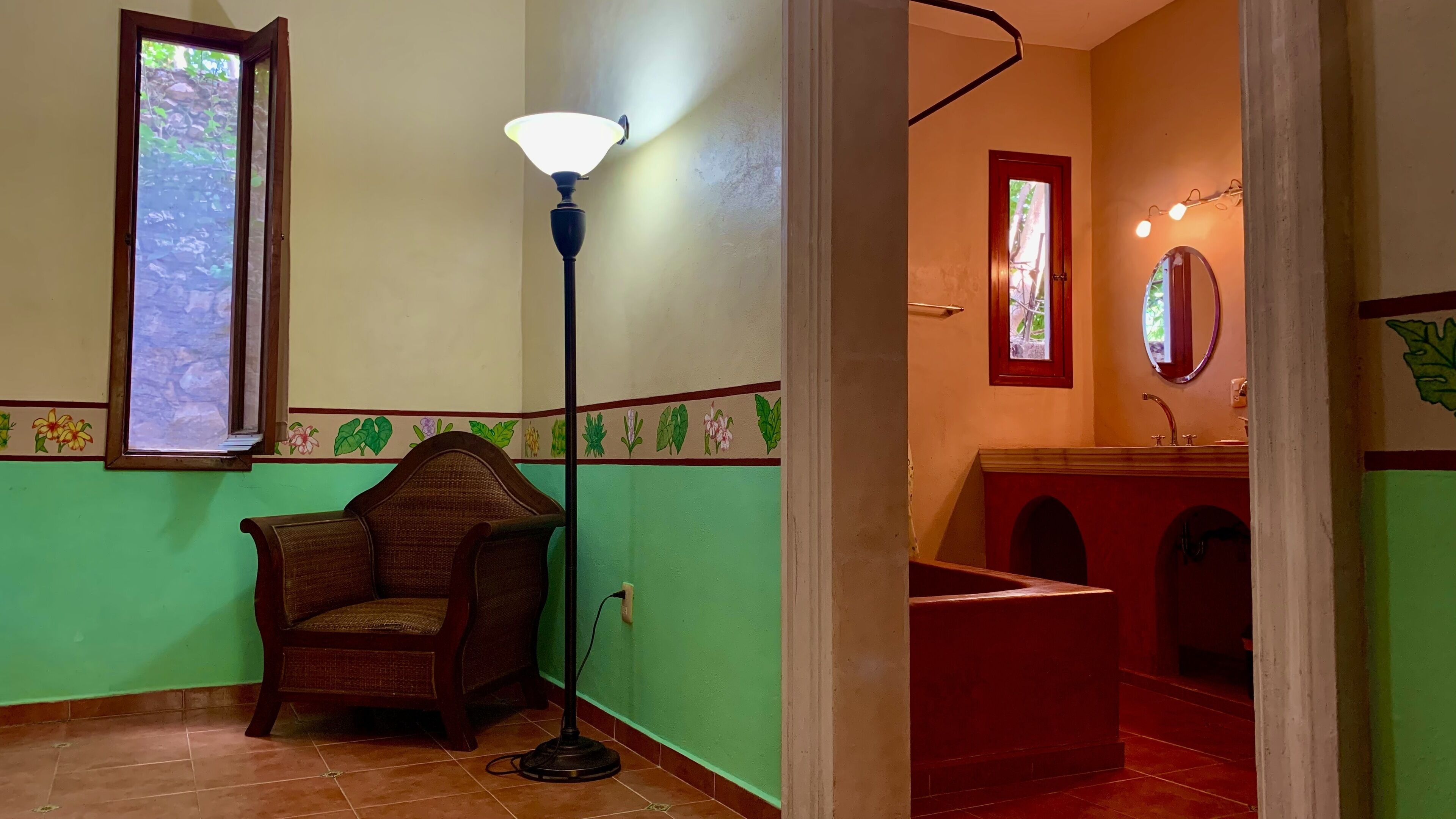 A room with a green wall, a wooden chair, a floor lamp, a window, a mirror, and a sink.