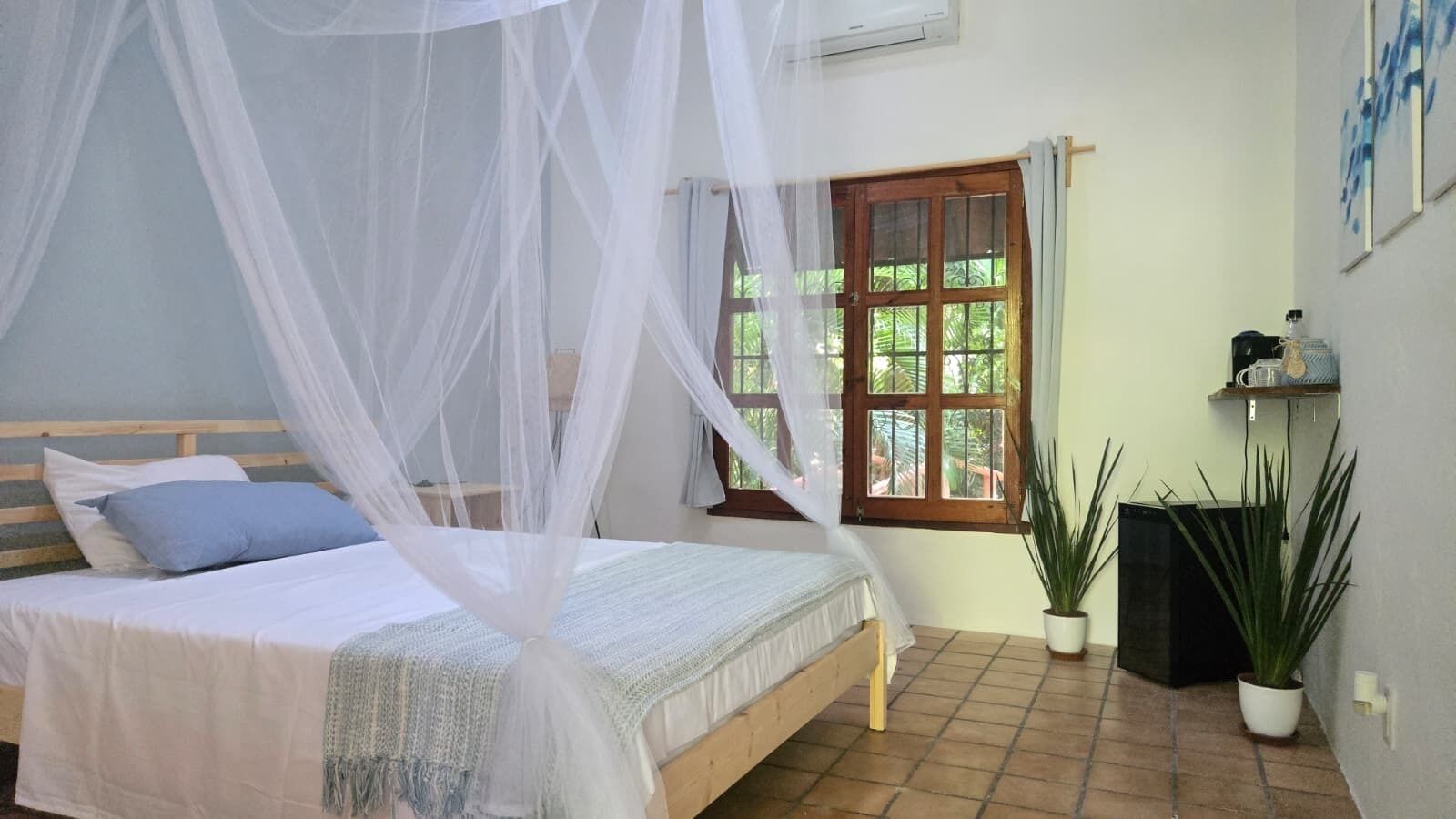 Un dormitorio con una cama bajo un dosel blanco, una ventana con vista a la vegetación y una planta en maceta en el suelo.