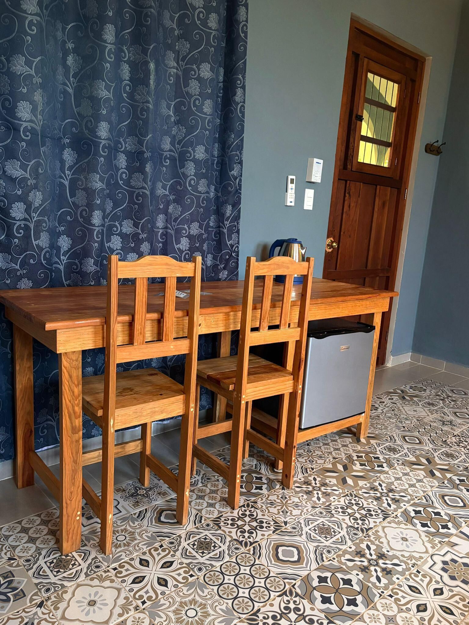 A room with a wooden table and chairs, a small refrigerator, a patterned curtain, a wooden door, and a tiled floor.