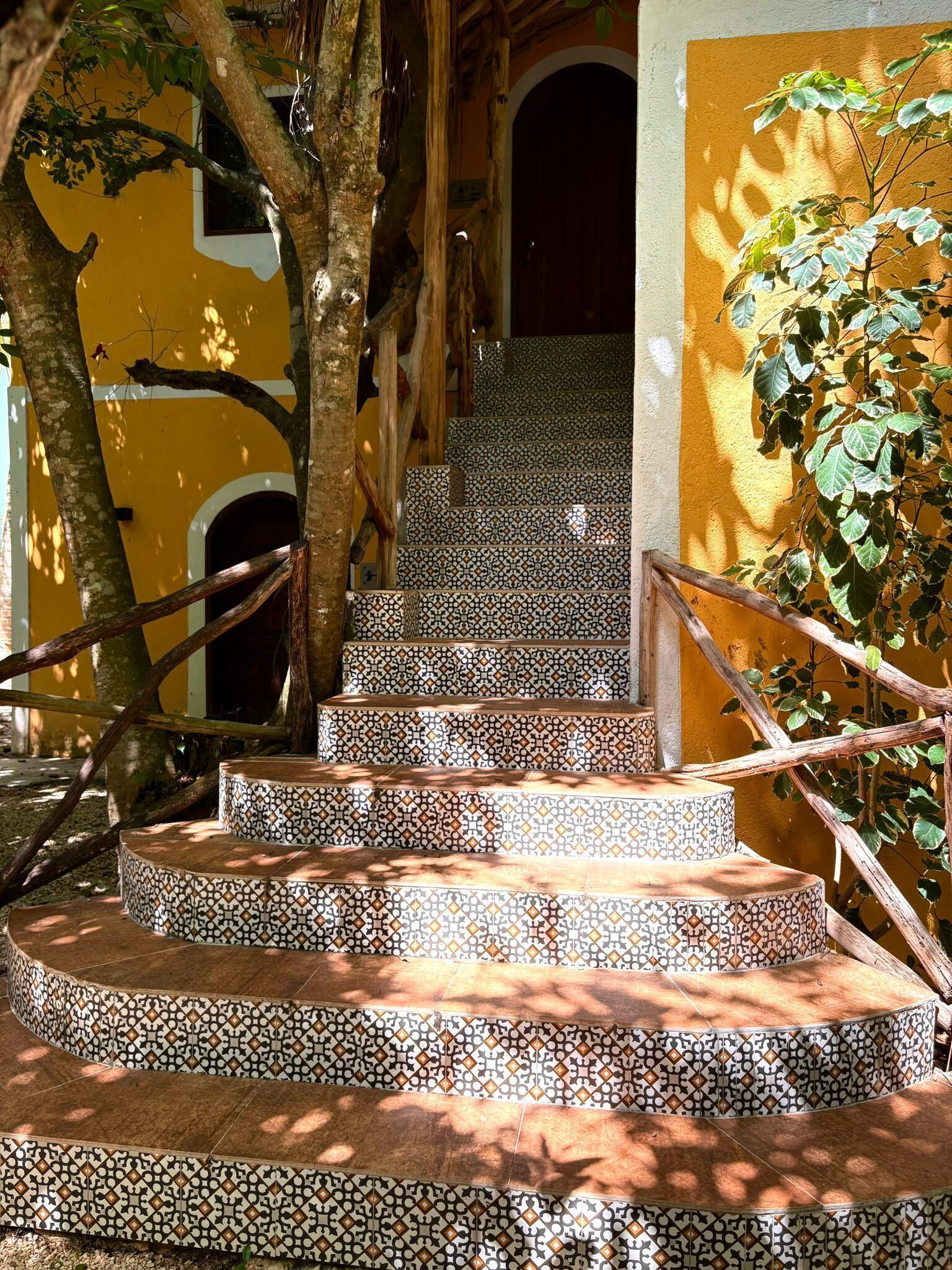 A set of outdoor stairs with patterned tiles leading up to a door, surrounded by trees and a yellow wall.
