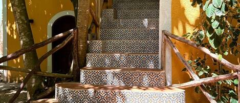 A set of outdoor stairs with patterned tiles leading up to a door, surrounded by trees and a yellow wall.