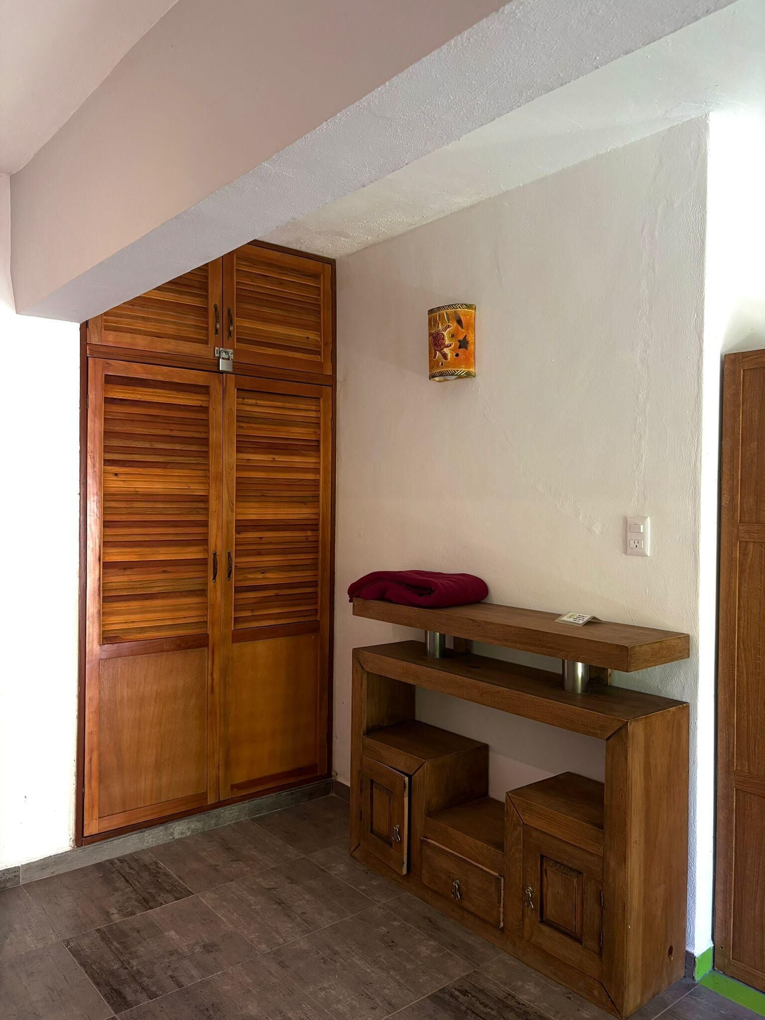 Una habitación con un armario de madera, un pequeño estante con un cojín rojo, y una lámpara de pared.