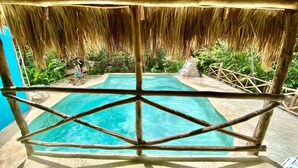 A pool with a thatched roof and wooden railing, surrounded by lush greenery.