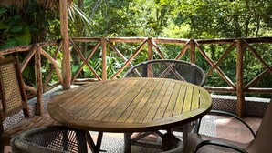A wooden table and chairs under a thatched roof, surrounded by a wooden fence with a view of lush greenery.