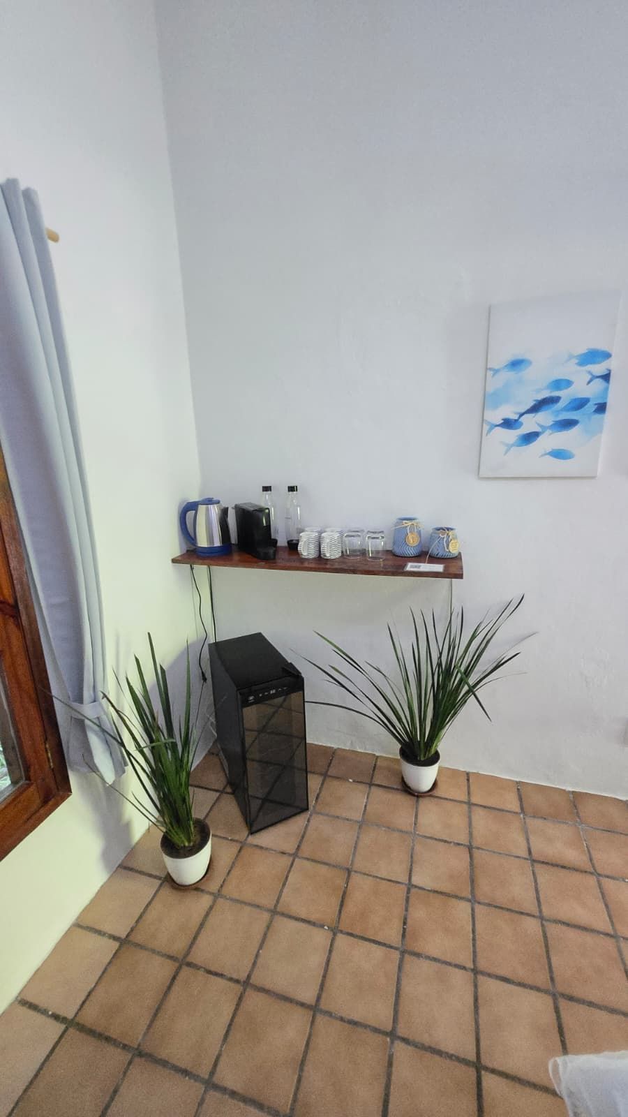 Una habitación de esquina con suelo de baldosas, un pequeño calentador negro, dos plantas en macetas, un estante con utensilios de cocina y una pared con un cuadro de un pez azul.