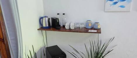 Una habitación de esquina con suelo de baldosas, un pequeño calentador negro, dos plantas en macetas, un estante con utensilios de cocina y una pared con un cuadro de un pez azul.