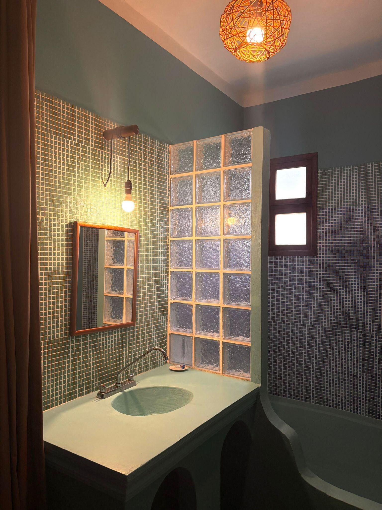 Un baño con pared de azulejos verdes, un espejo, un lavamanos y una ventana con paneles de vidrio esmerilado.