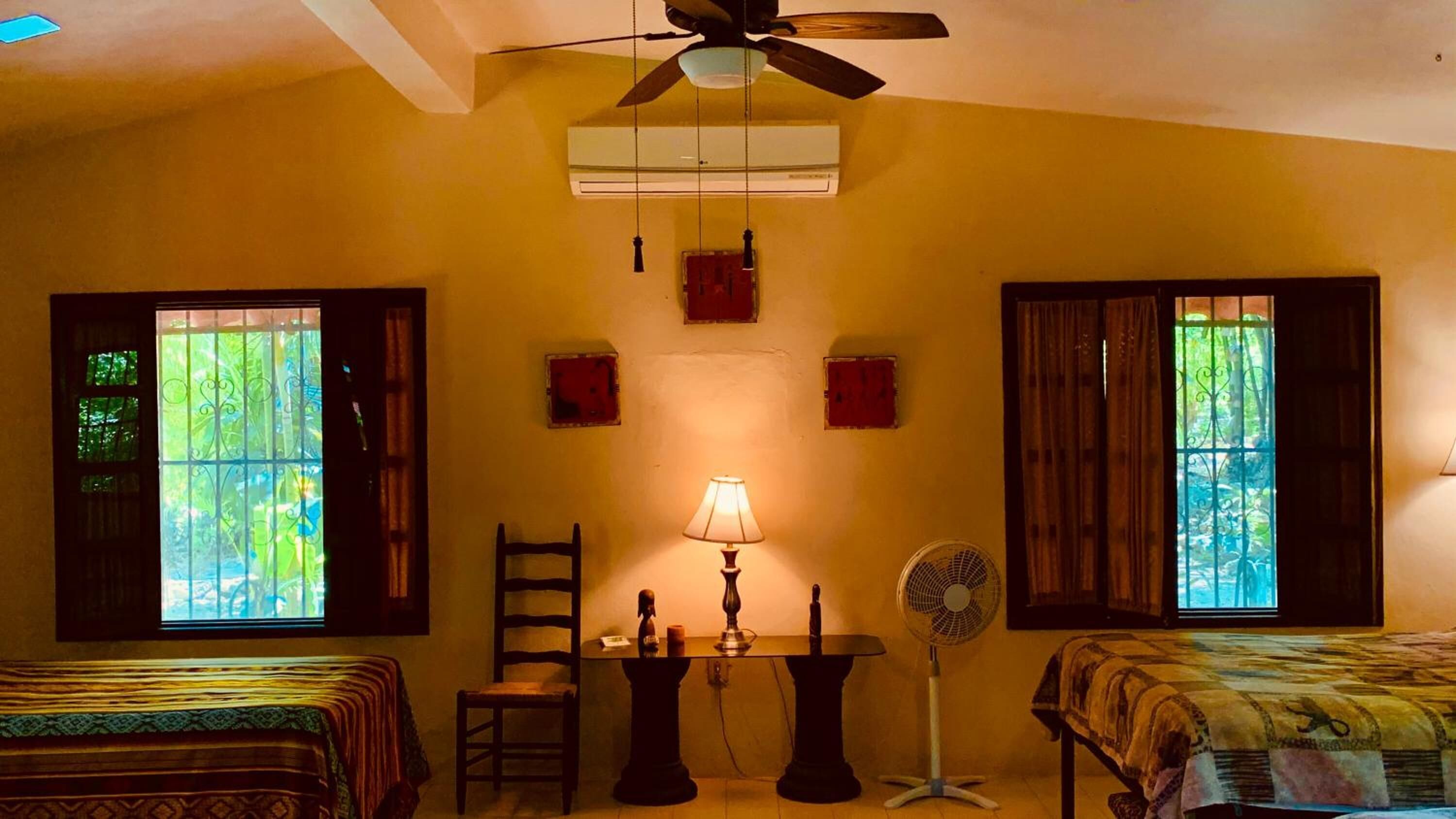 A room with two beds, a table with a lamp, a chair, a standing fan, and an air conditioning unit.