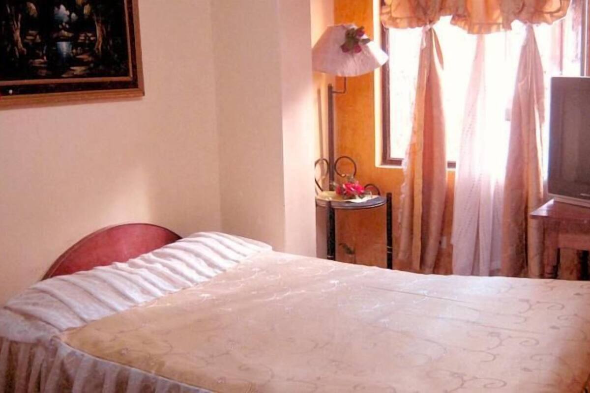 A bedroom with a bed, a chair, a table with a lamp, and a television.