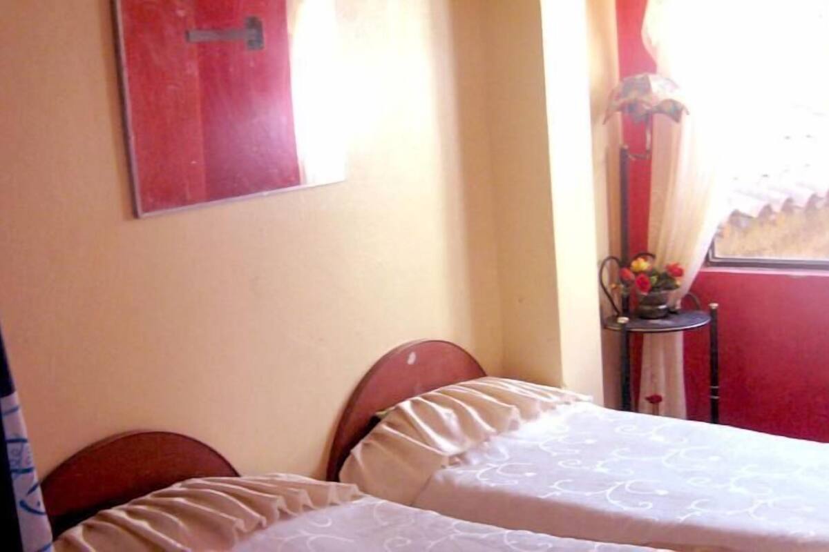 A room with two beds, a red door, a window with a curtain, and a small table with flowers.