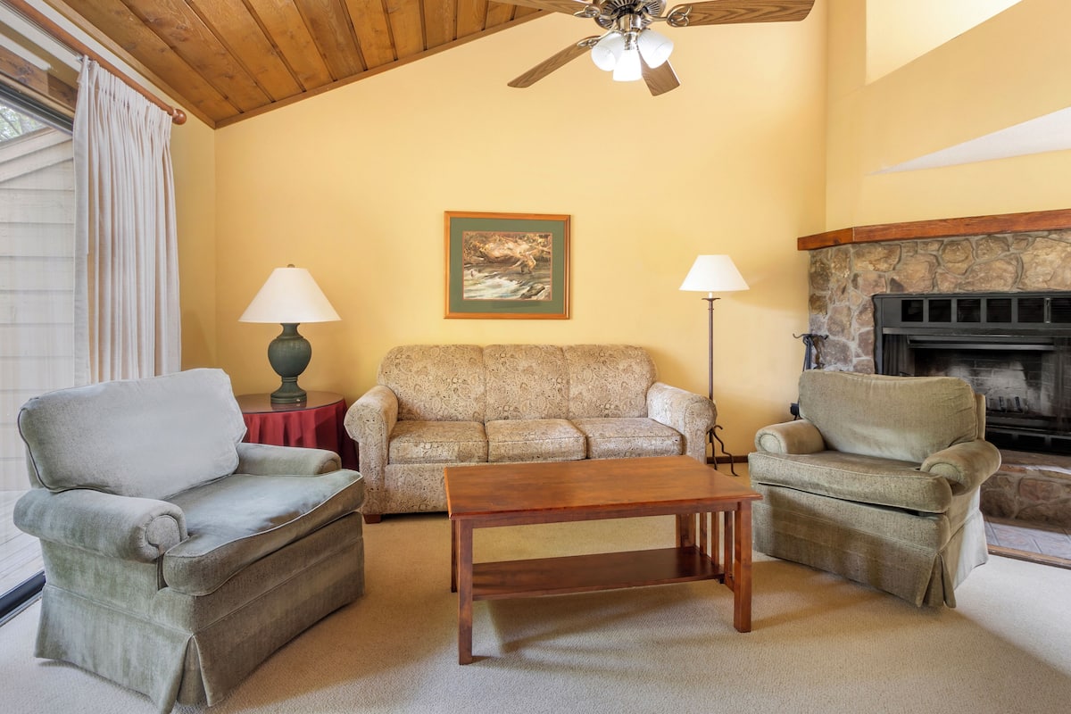 A cozy living room with a wooden ceiling, a stone fireplace, and two armchairs.
