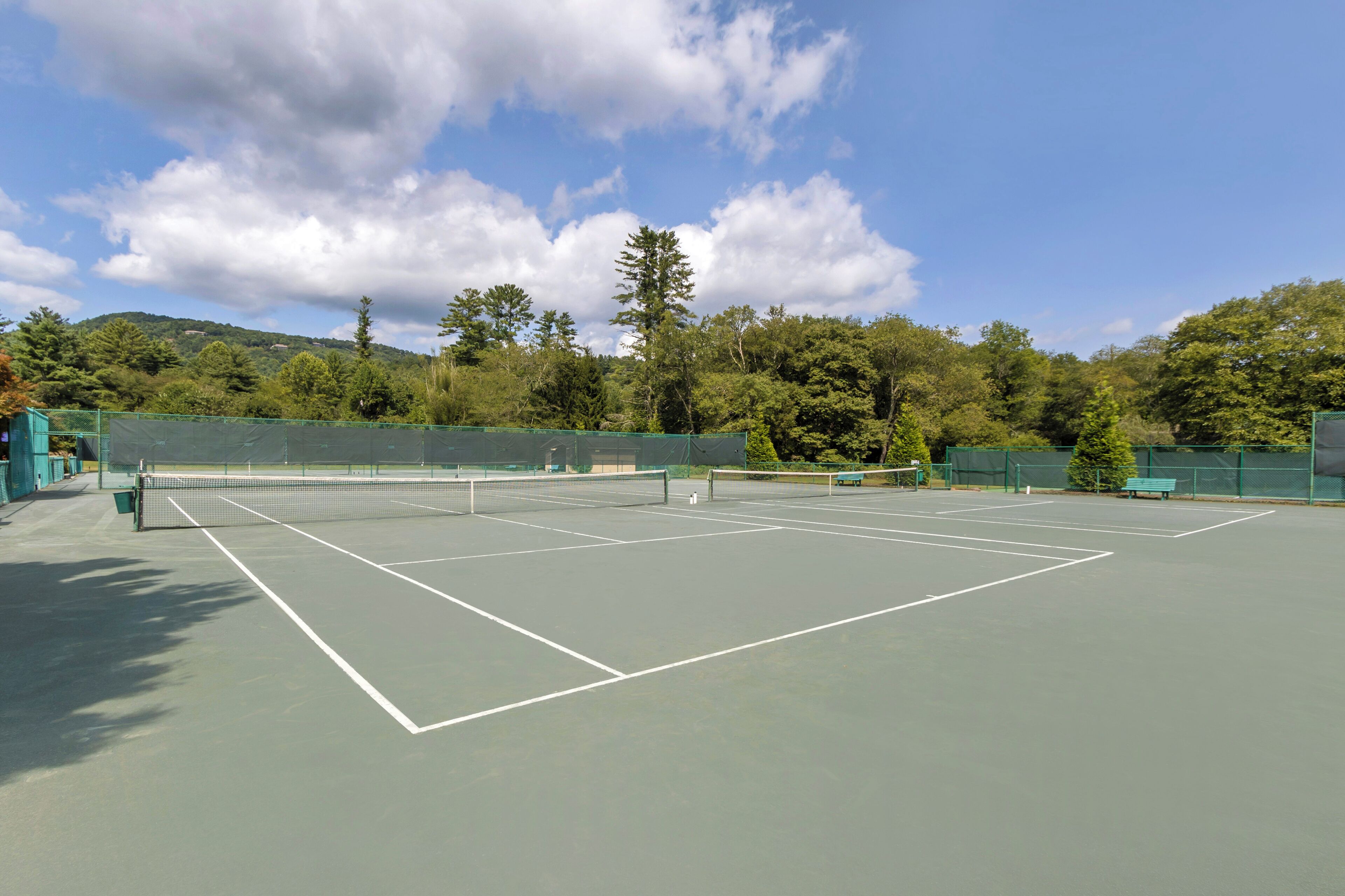 An outdoor tennis court with a net, surrounded by green fencing and trees.