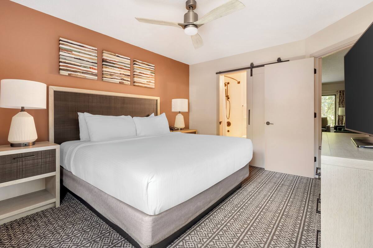 A hotel room with a large bed, two bedside lamps, a ceiling fan, a shower area, and a patterned floor.