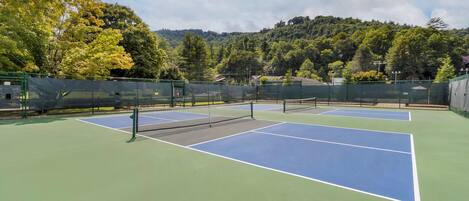 Un court de tennis avec un filet au centre, entouré d’une clôture verte et avec des arbres en arrière-plan.