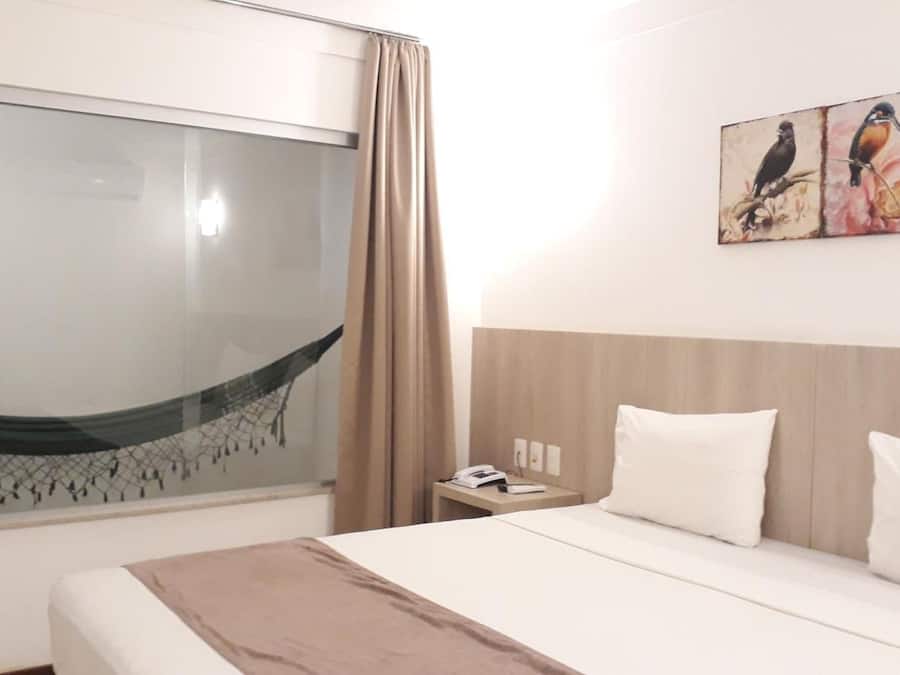 A hotel room with a bed, a nightstand, a painting of birds, a window with a view, and a curtain.