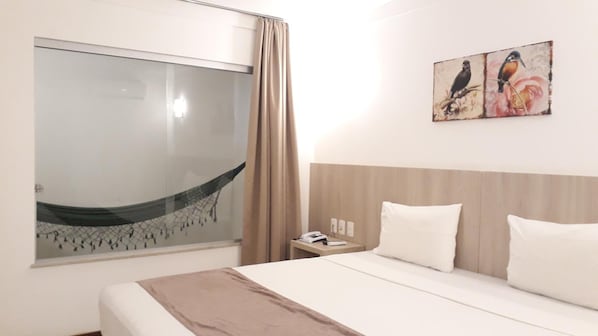 A hotel room with a bed, a nightstand, a painting of birds, a window with a view, and a curtain.